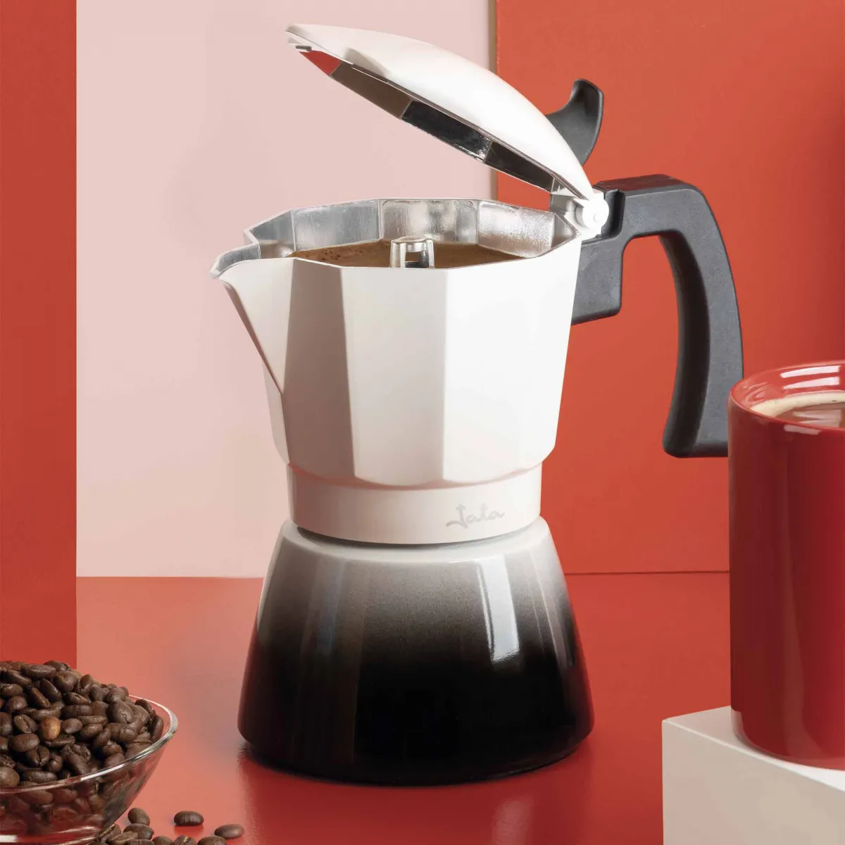 Una cafetera blanca y negra con granos de café y una taza roja.