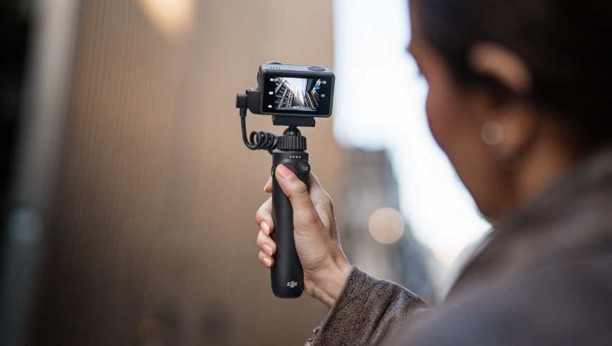 Osoba trzymająca czarną kamerę sportową na uchwycie, filmująca miasto.