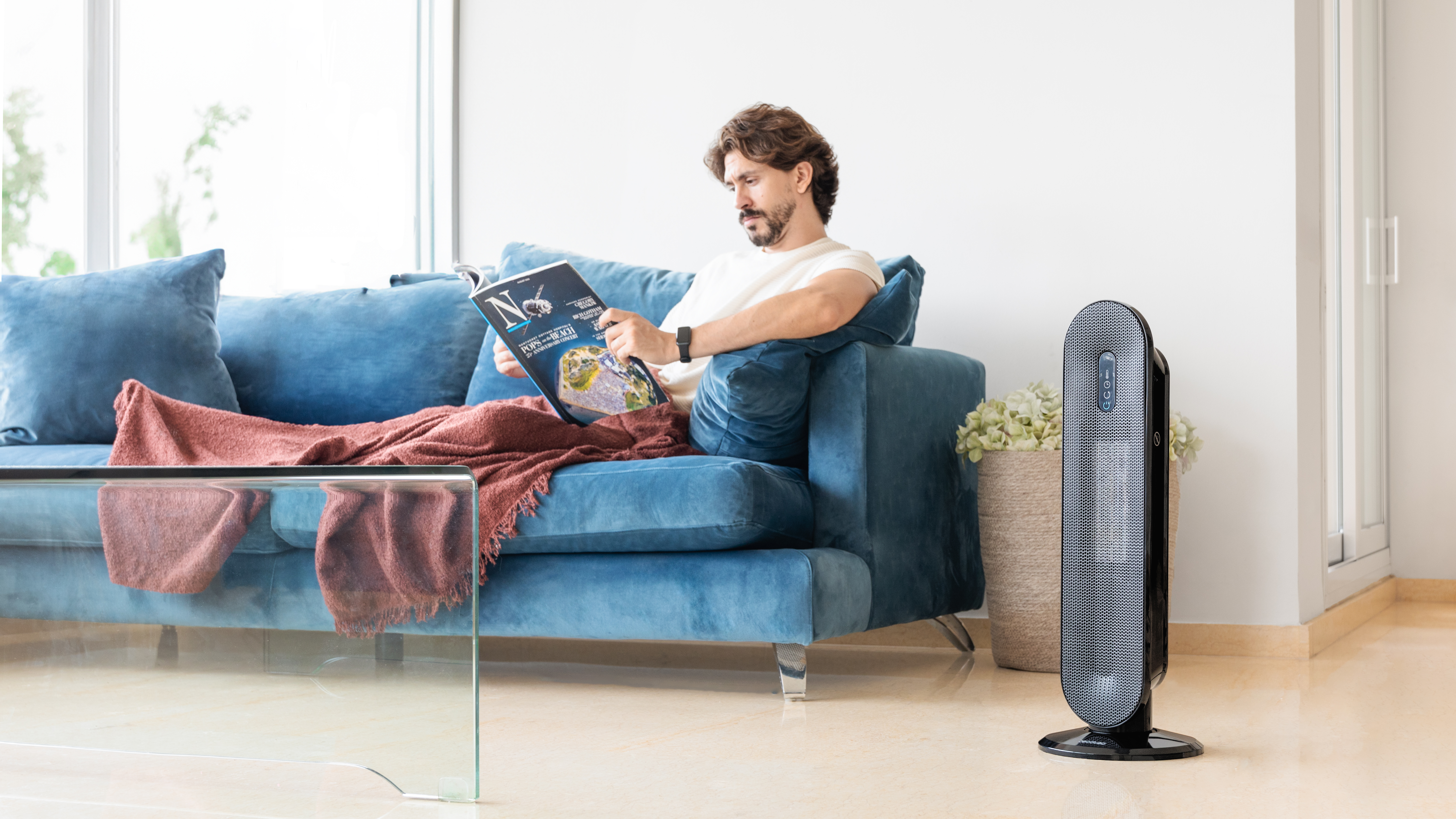 Hombre sentado en un sofá azul leyendo una revista, con un calefactor y una mesa de cristal cerca.