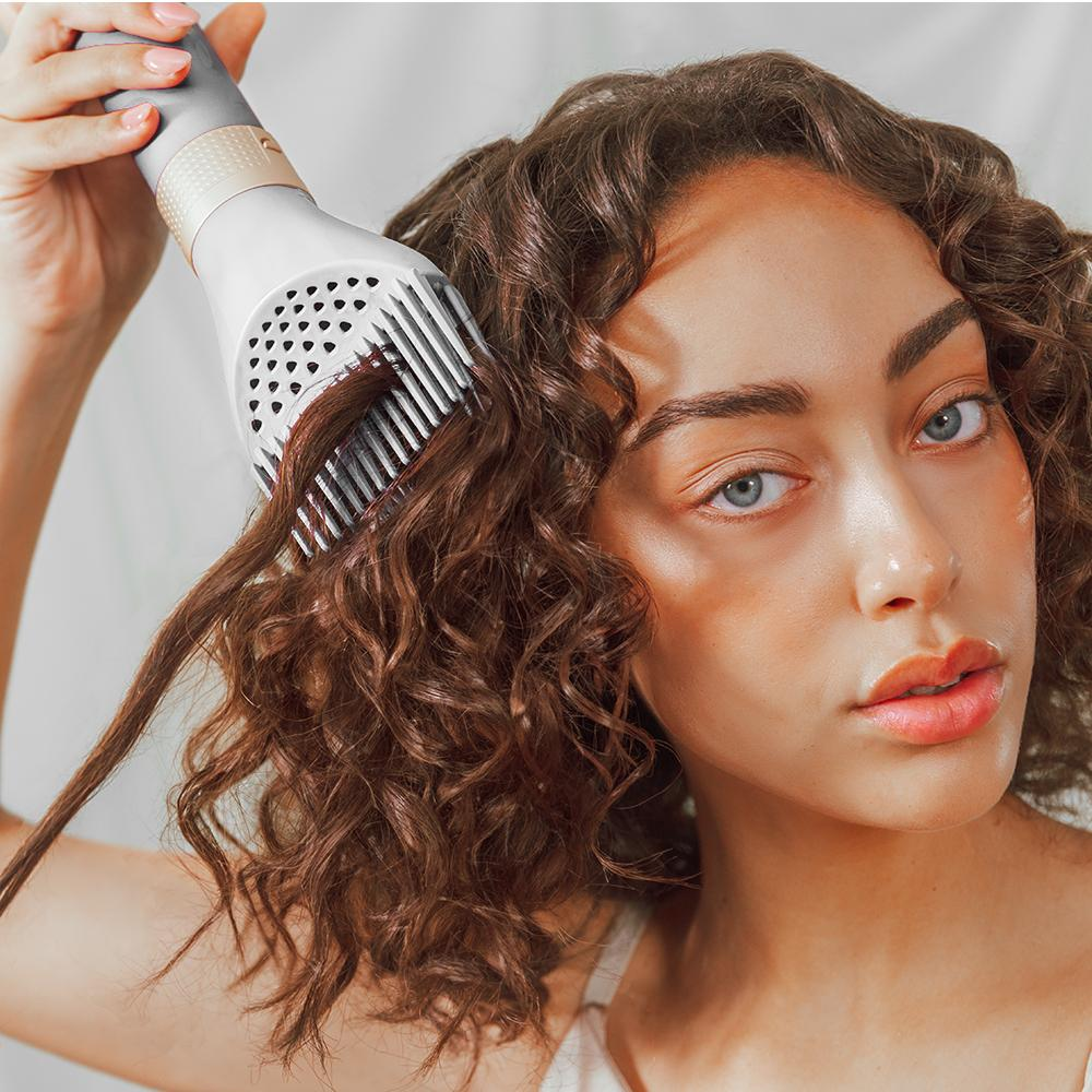 Una mujer usa un secador de pelo con un accesorio de peine para secar el cabello rizado. Mira a la cámara.