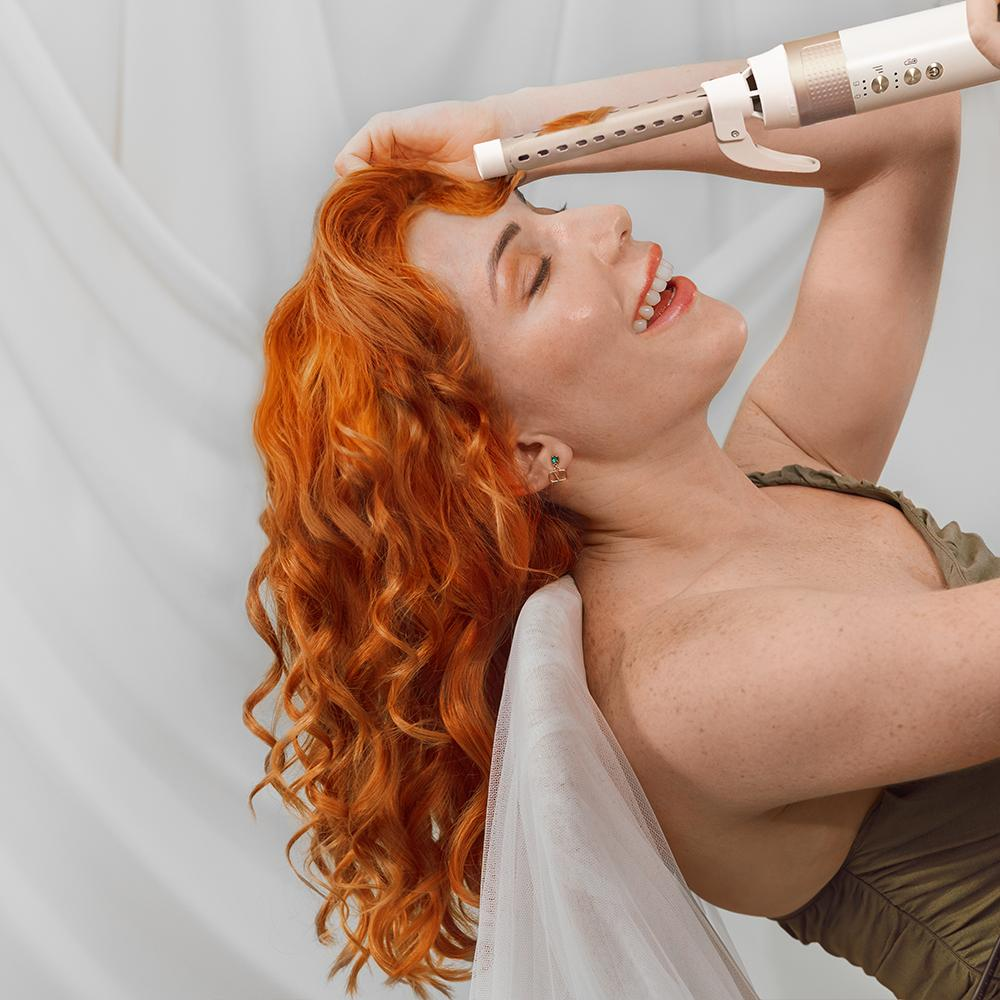 Una mujer con cabello rizado rojo sonríe mientras usa una herramienta para rizar el cabello.
