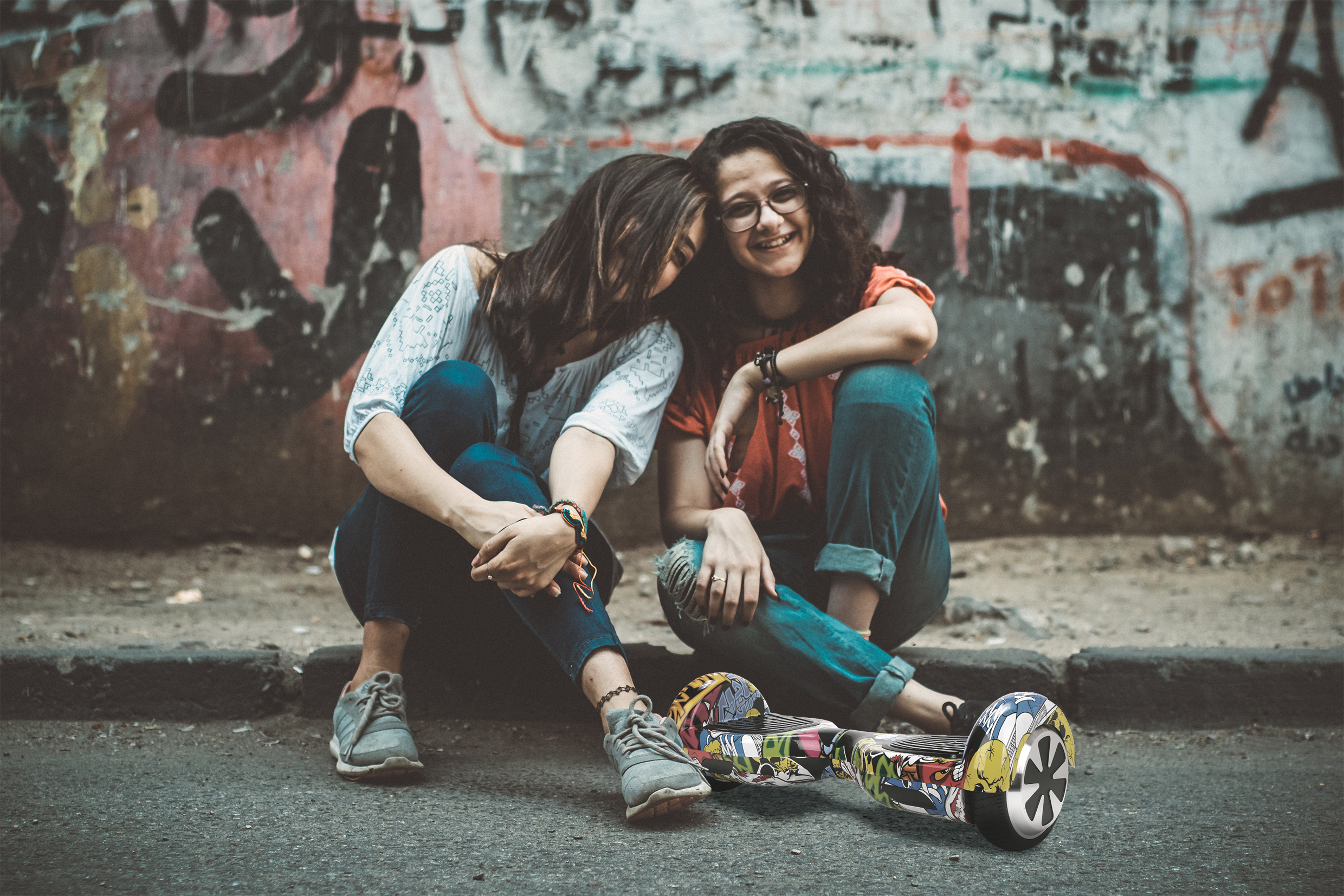 Zwei Frauen sitzen, eine lehnt sich an die andere, mit einem Hoverboard im Vordergrund.