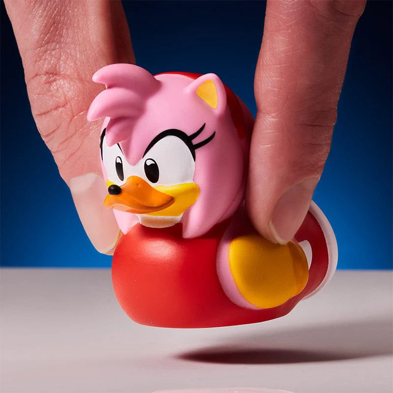 Kezek egy Amy Rose gumikacsát tartanak. Rózsaszín haj, piros test és sárga részletek láthatók.