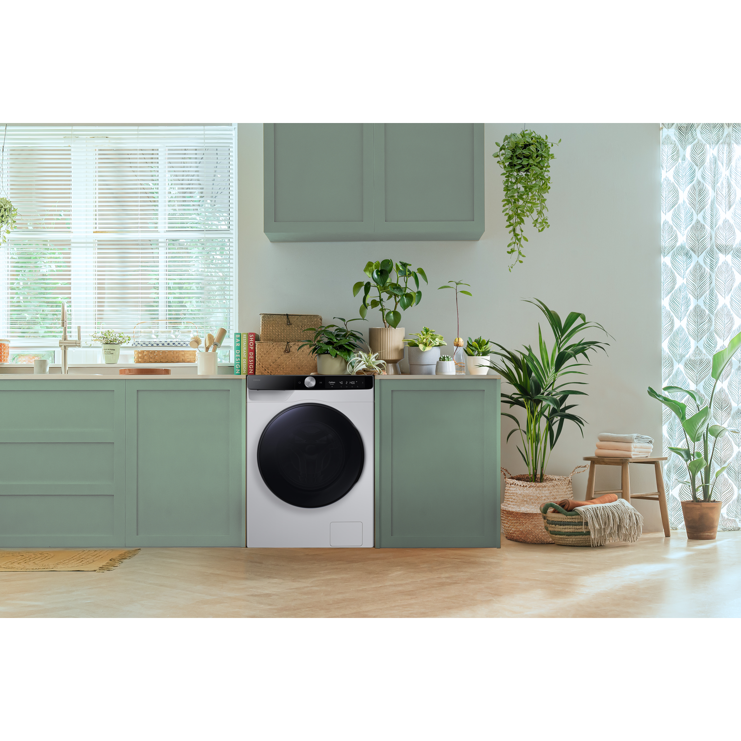 Een wasmachine in een keukenomgeving, naast groene kasten en planten.
