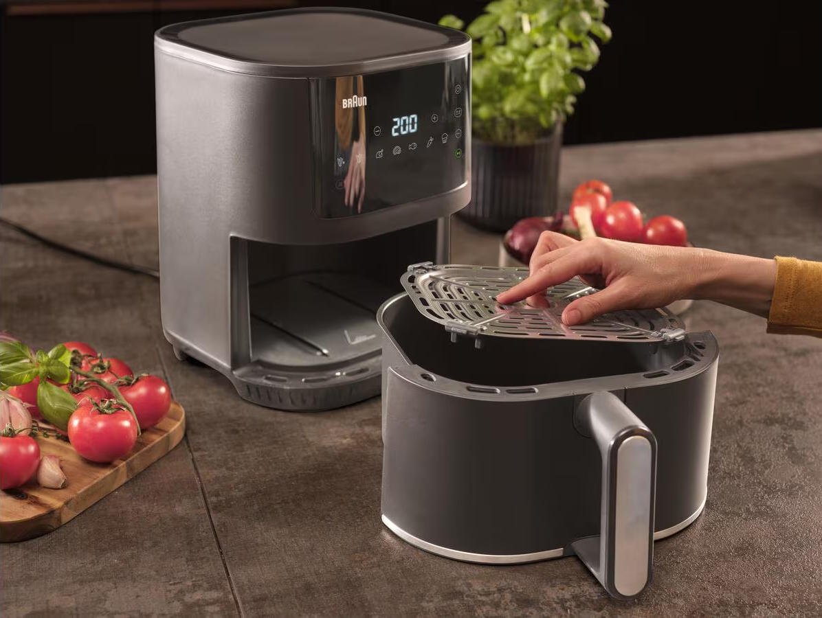 Una friggitrice ad aria con una mano che inserisce una griglia all'interno. Ci sono pomodori e una pianta nelle vicinanze.