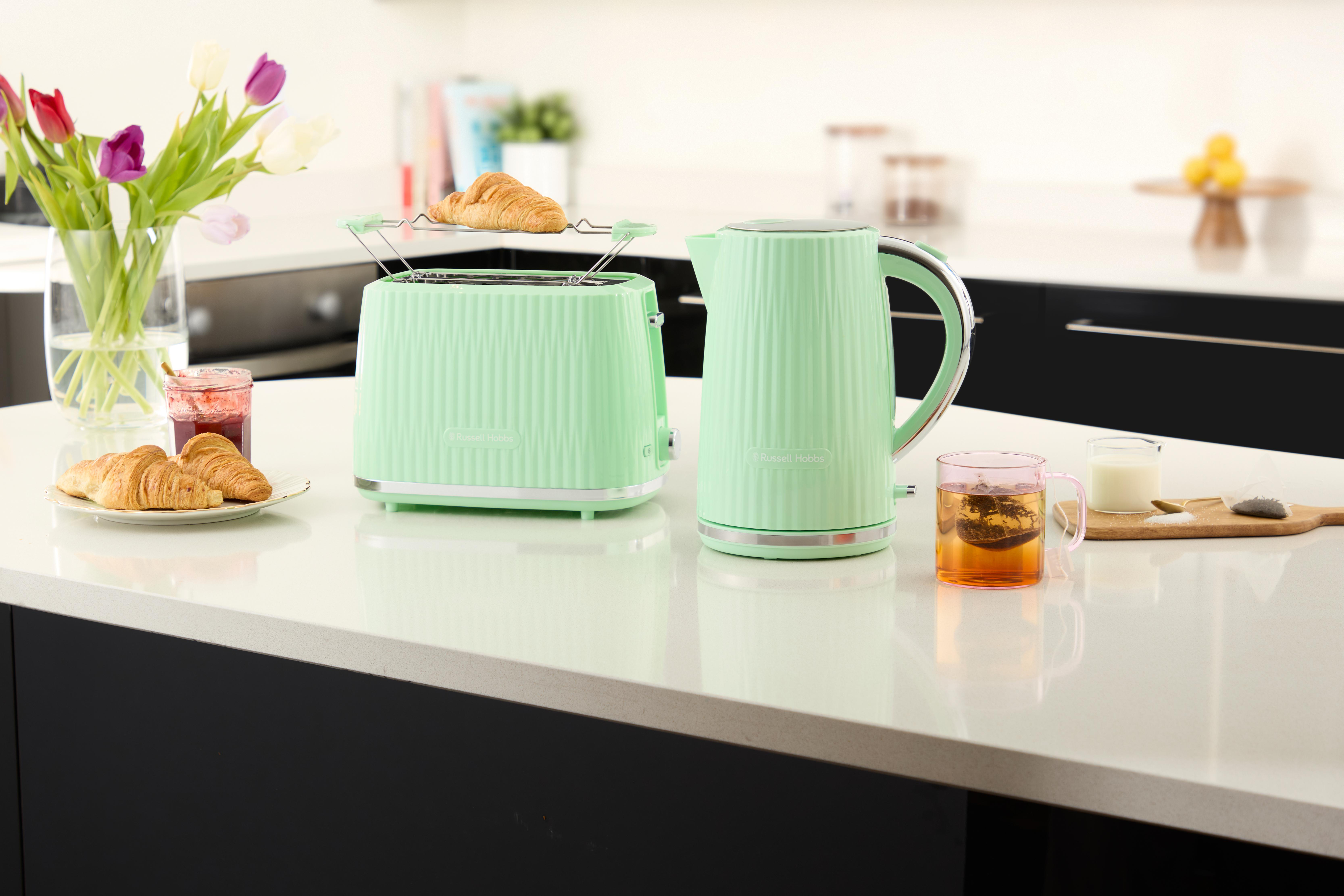 Eine Küchenszene mit Toaster, Wasserkocher und Essen auf einer Arbeitsplatte. Blumen in einer Vase. Hintergrund schwarz.