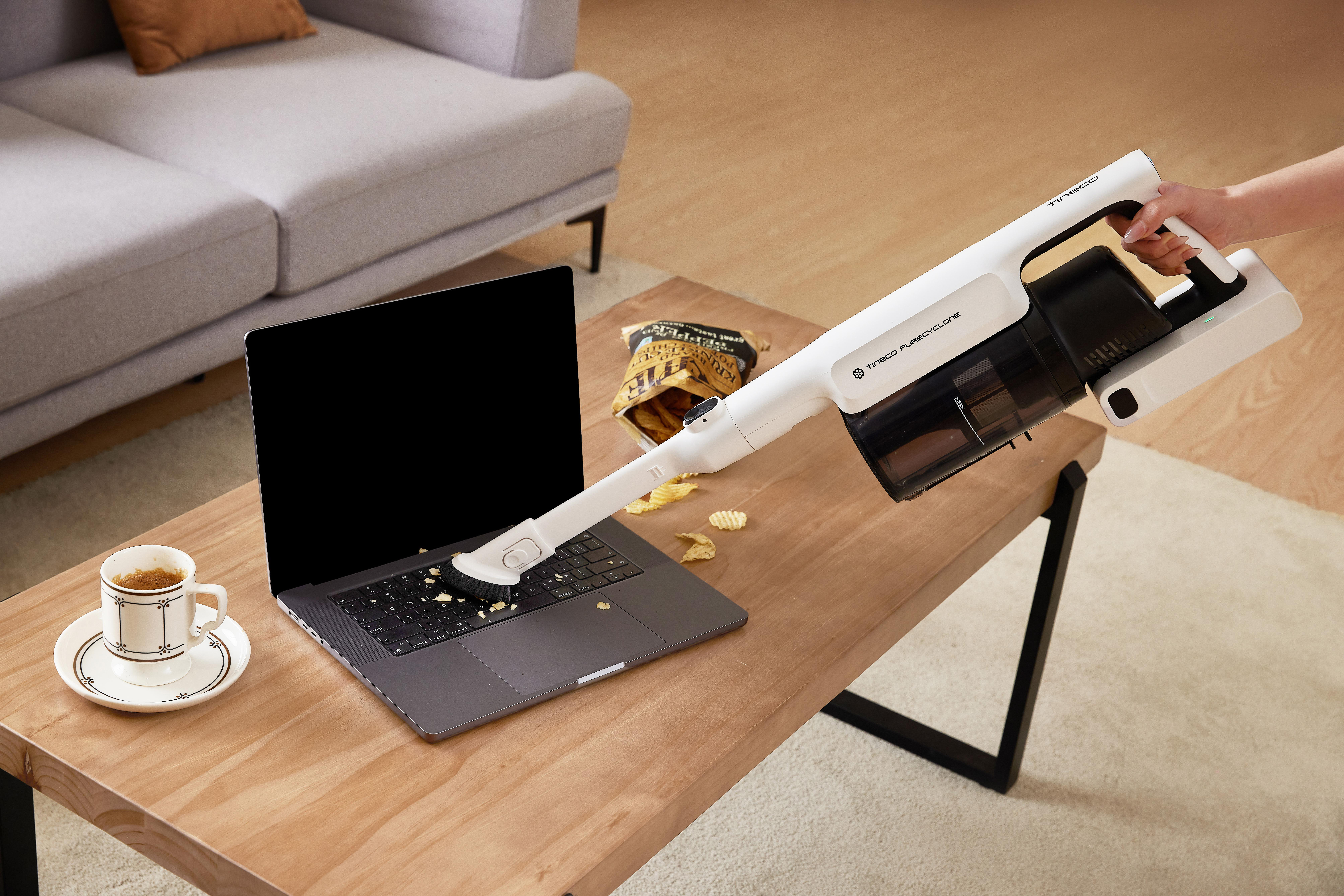 Eine Person saugt mit einem weißen Staubsauger die Tastatur eines Laptops auf einem Tisch ab.