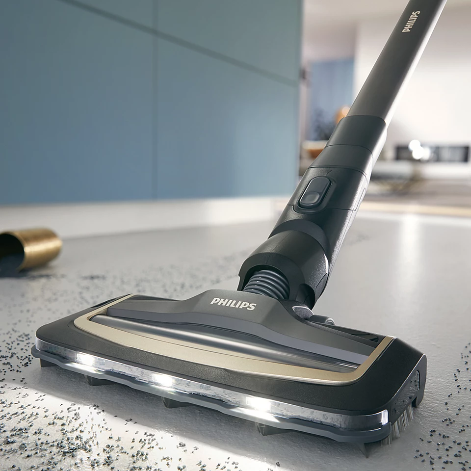 Un aspirapolvere Philips nero e grigio pulisce un pavimento con detriti sparsi.