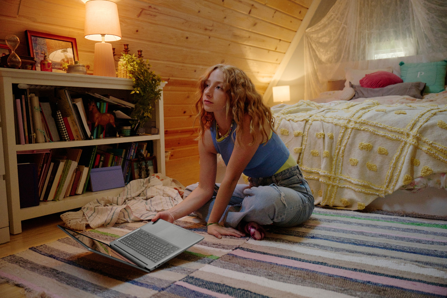 Een jonge vrouw met krullend haar zit op een kleed en gebruikt een laptop in een gezellige slaapkamer.