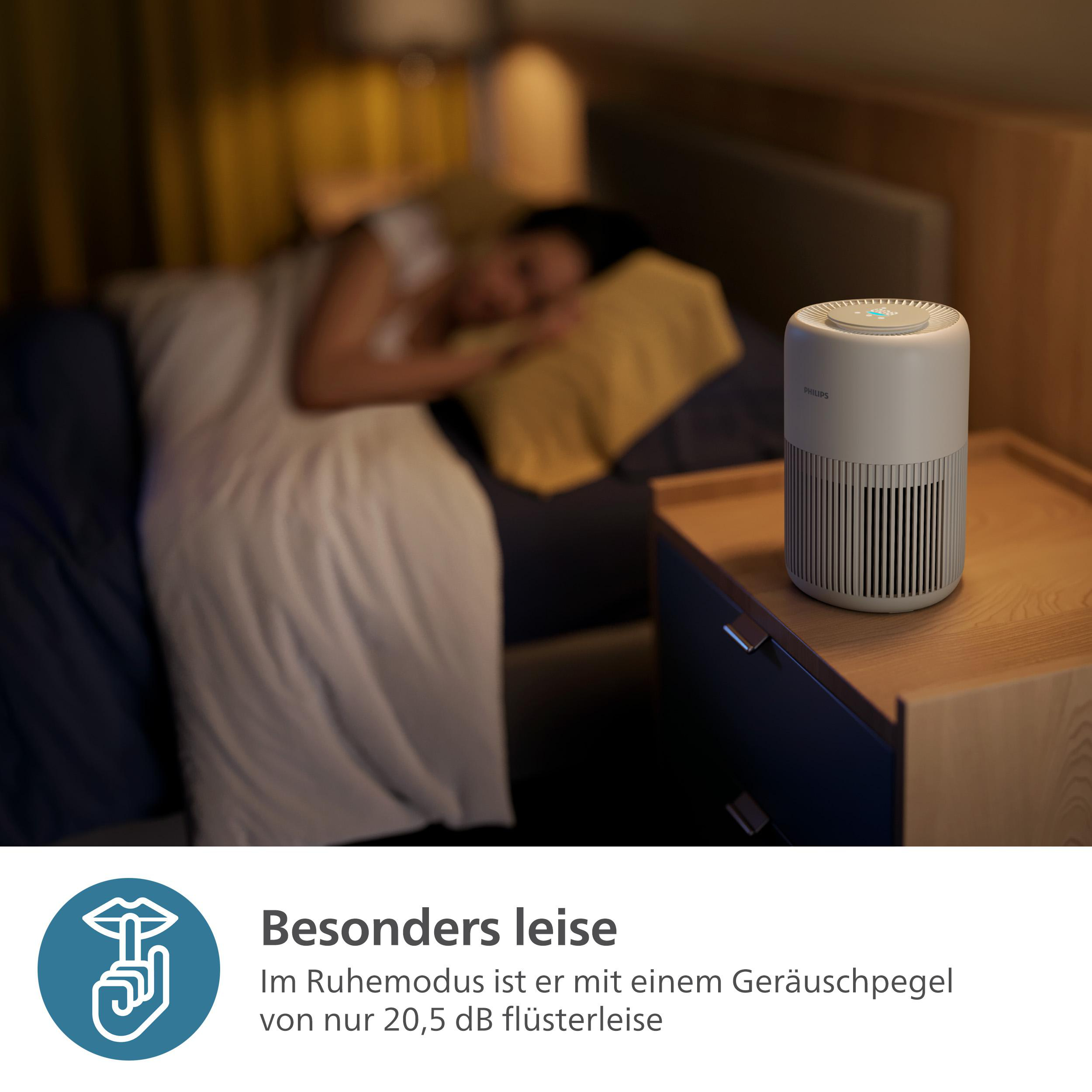 Luftreiniger auf einem Nachttisch neben einem Bett. Der Hintergrund ist unscharf.