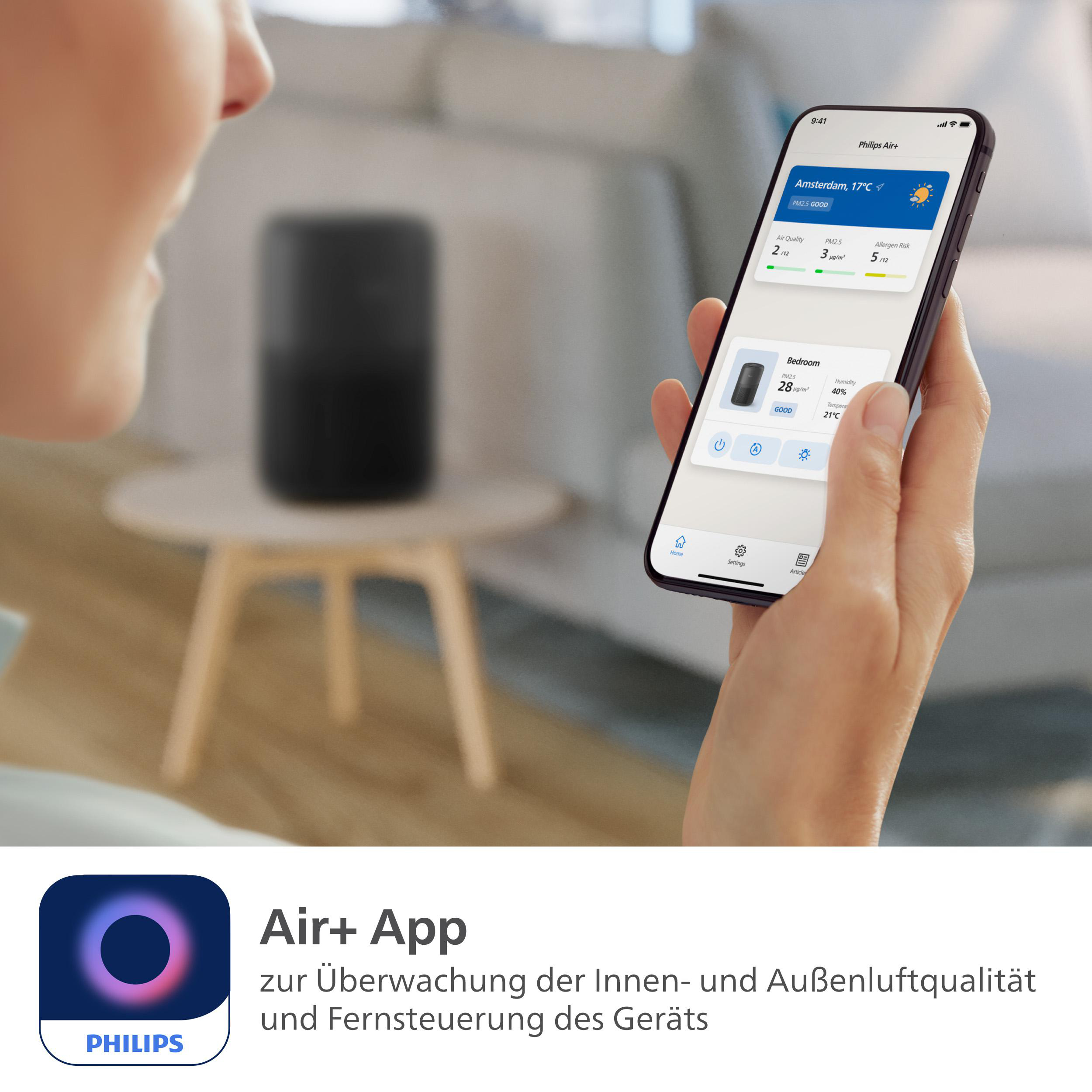 Frau hält Telefon mit Luftqualitäts-App, in der Nähe eines schwarzen Luftreinigers und eines Tisches.