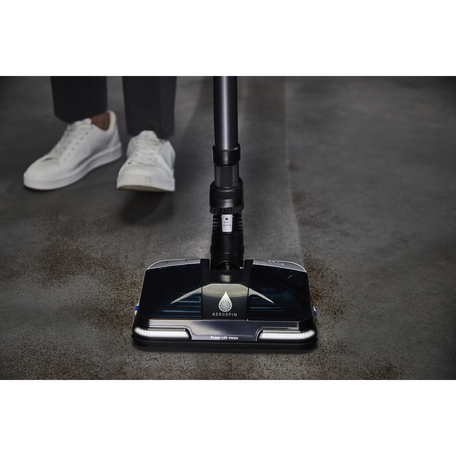 Vacuum cleaner head on a concrete floor. Person's legs and feet in white sneakers in the background.