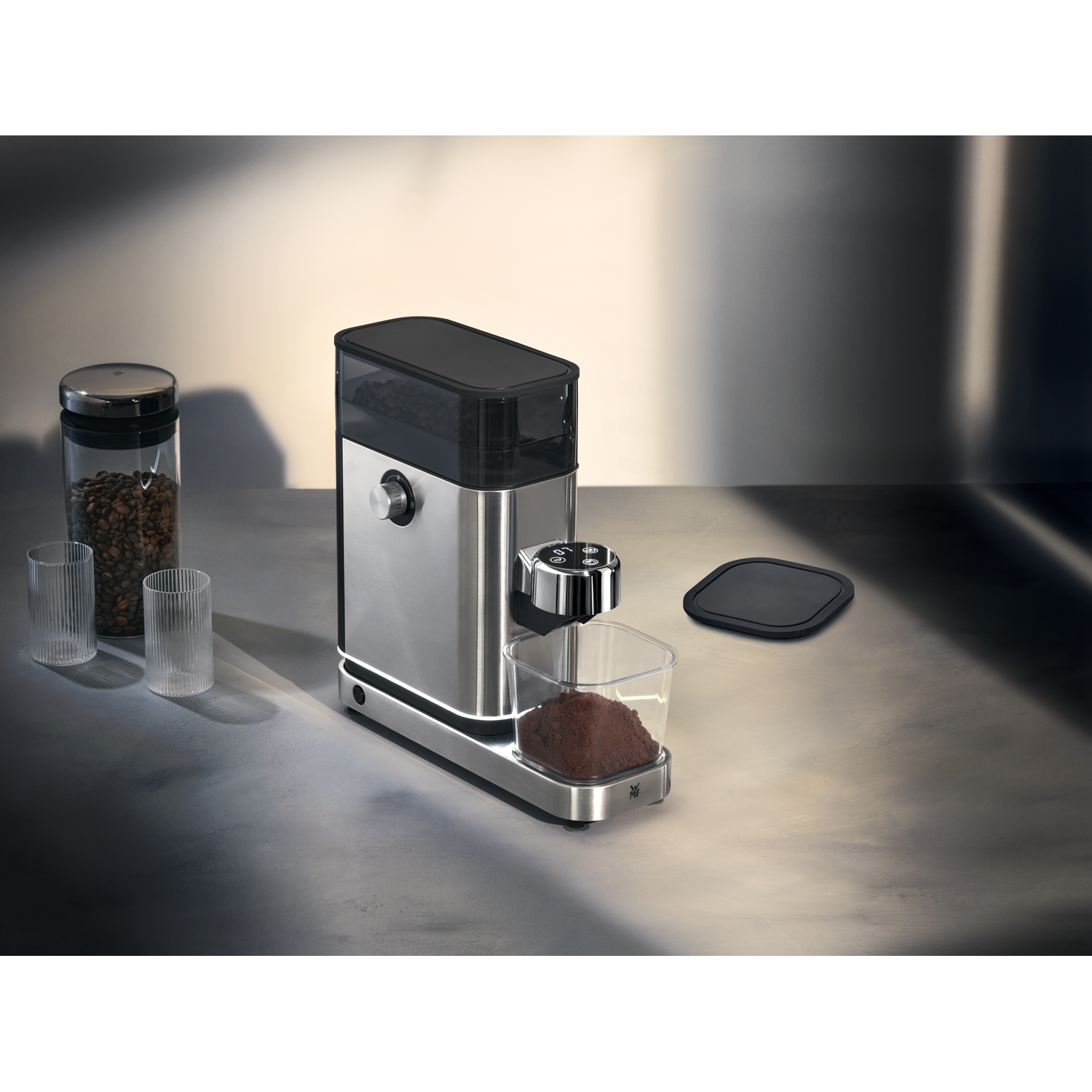 Stainless steel coffee grinder with a container of coffee beans and two glasses on the left.