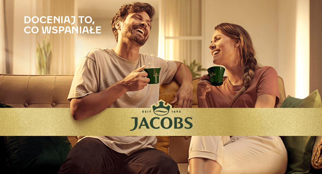 Uśmiechnięta para trzymająca filiżanki kawy Jacobs, z logo Jacobs, kanapą i ciepłym otoczeniem.