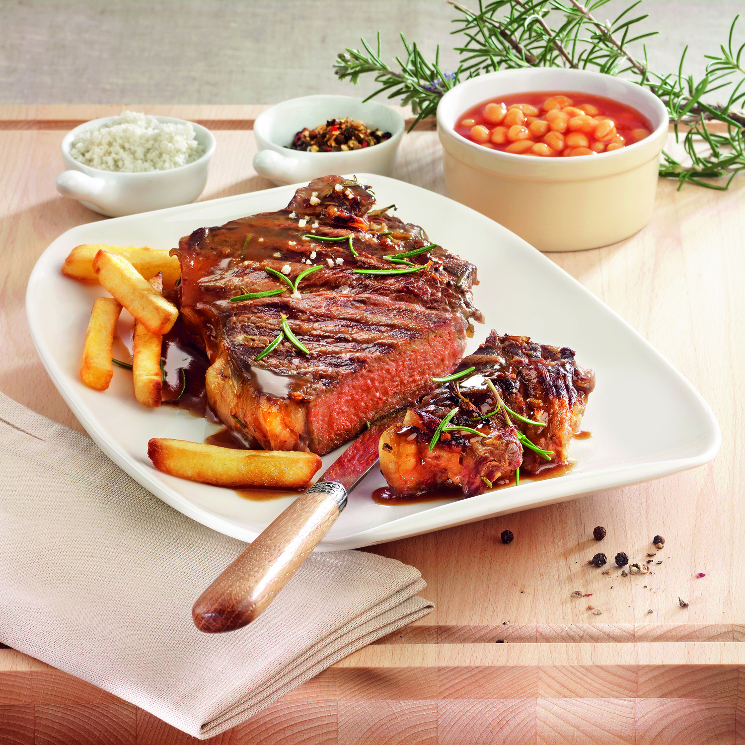 Steak sur une assiette blanche avec frites et haricots. Romarin et sel à côté, table en bois.