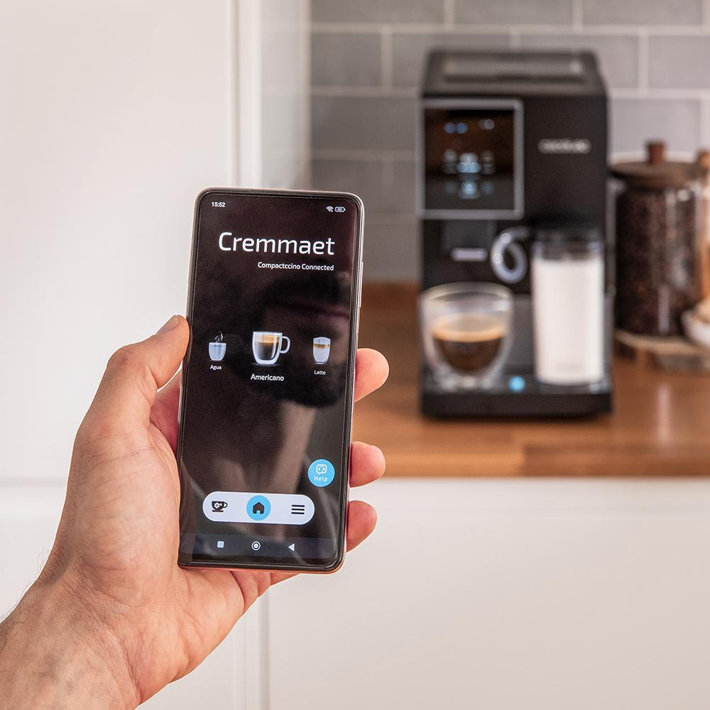 Mano sosteniendo teléfono muestra app cafetera, cafetera al fondo.