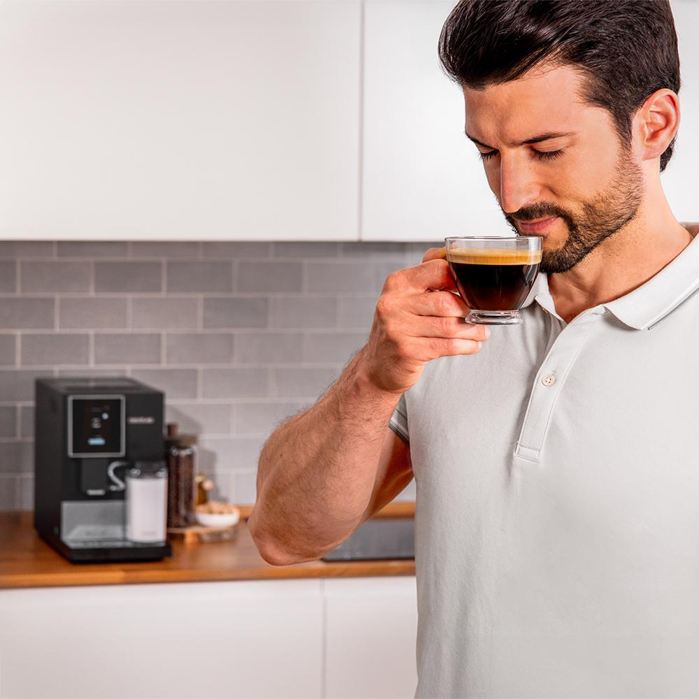 Un hombre oliendo café de una taza de vidrio transparente. Una cafetera está en la encimera.