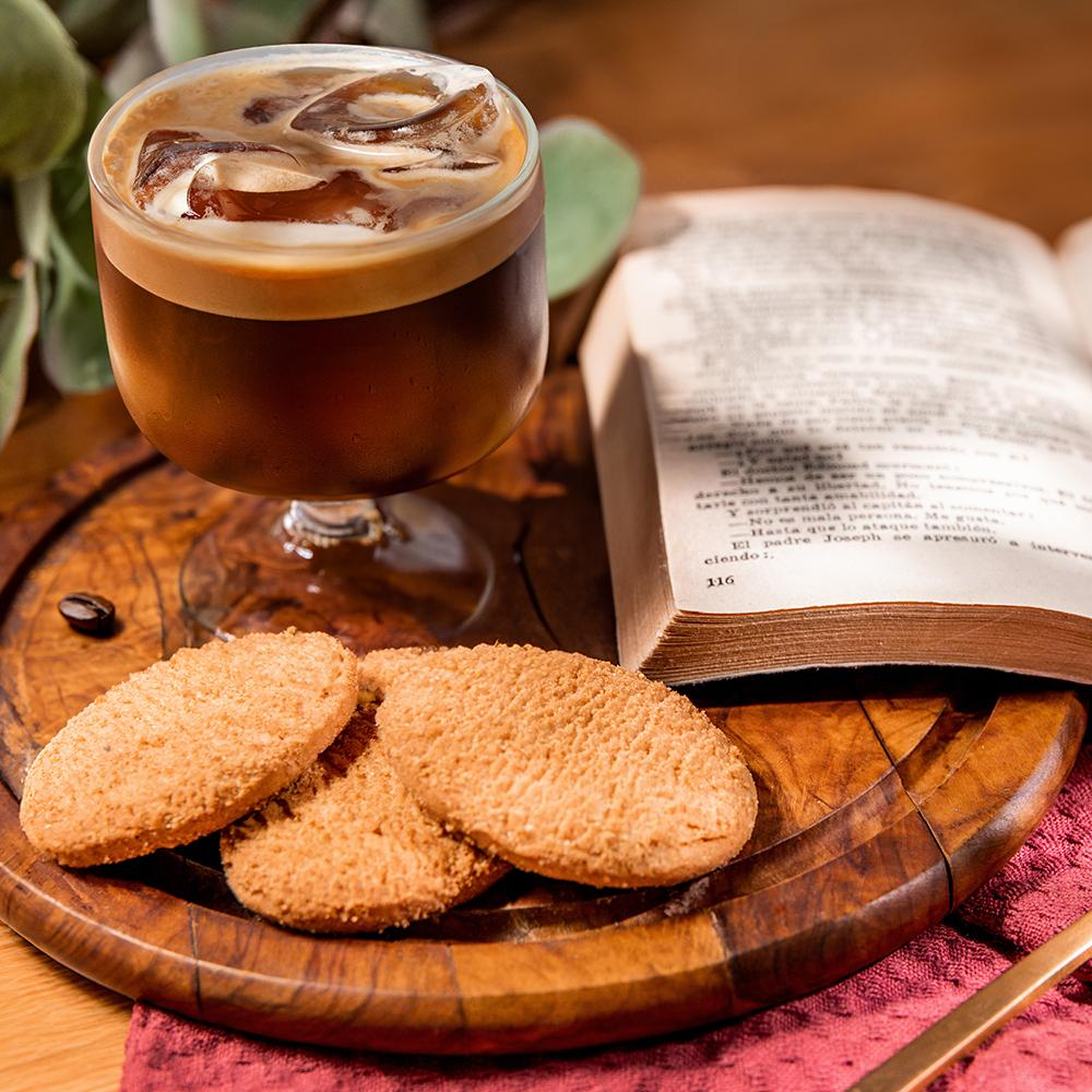 Bebida de café en un vaso, con galletas y un libro en una bandeja de madera. Fondo oscuro.