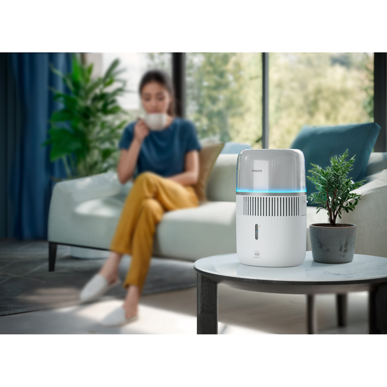 A woman drinking tea by a window, near a purifier and a potted plant.