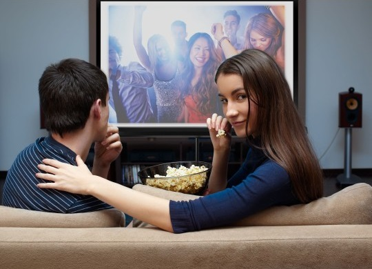 Para ogląda telewizję, jedząc popcorn. Na ekranie scena z imprezy.
