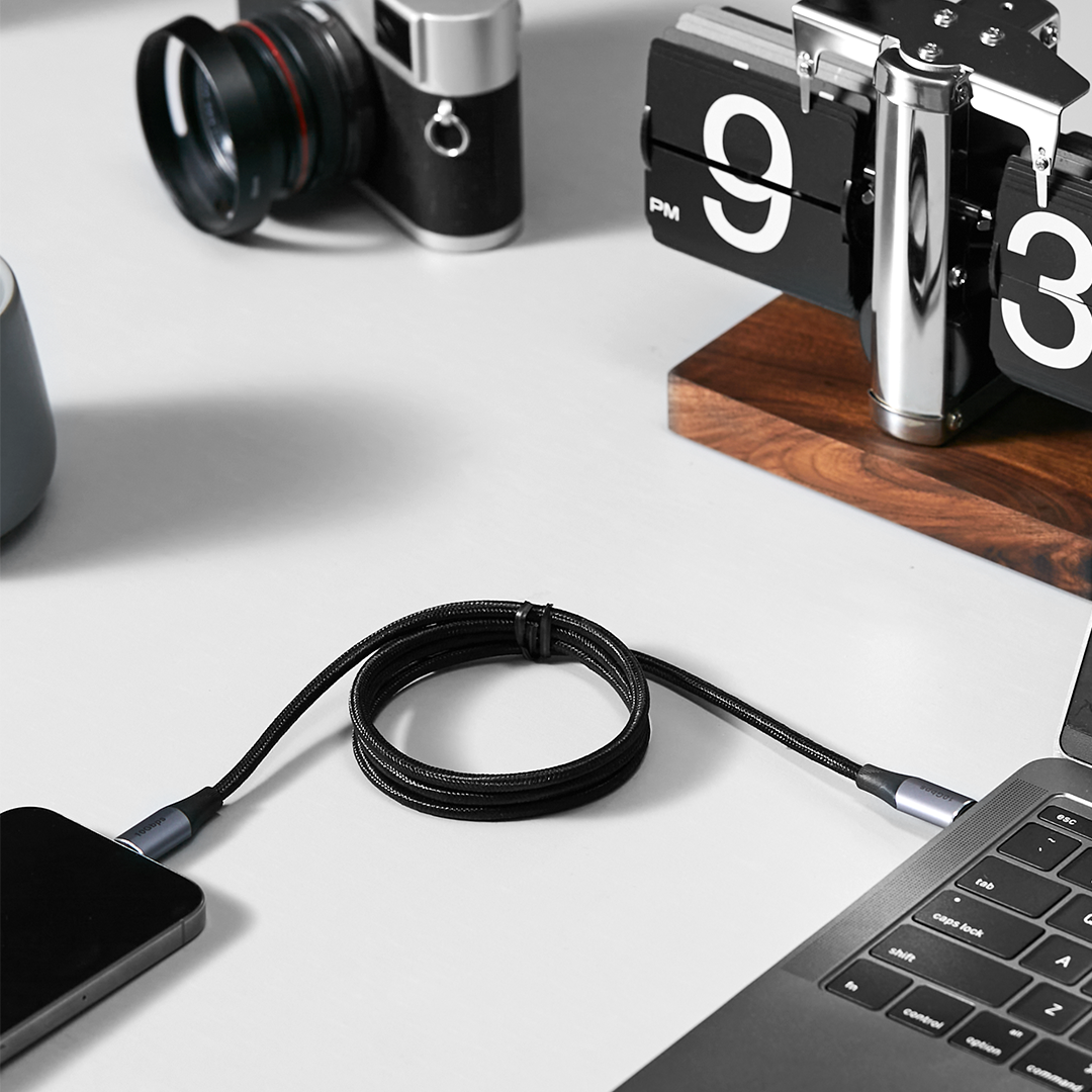 Telefon ve dizüstü bilgisayarı bağlayan siyah bir USB-C kablosu. Masada siyah bir kamera ve saat.