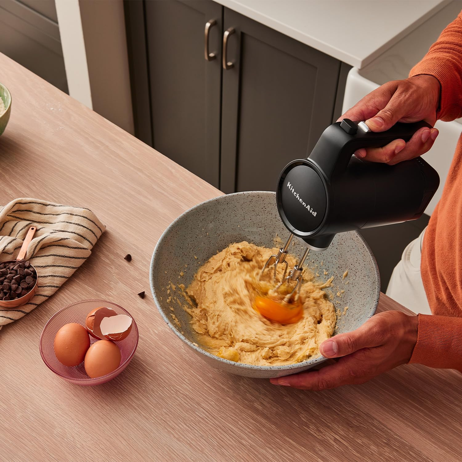 Egy személy tésztát kever egy tálban egy KitchenAid kézi mixerrel, tojásokkal és csokidarabokkal a közelben.