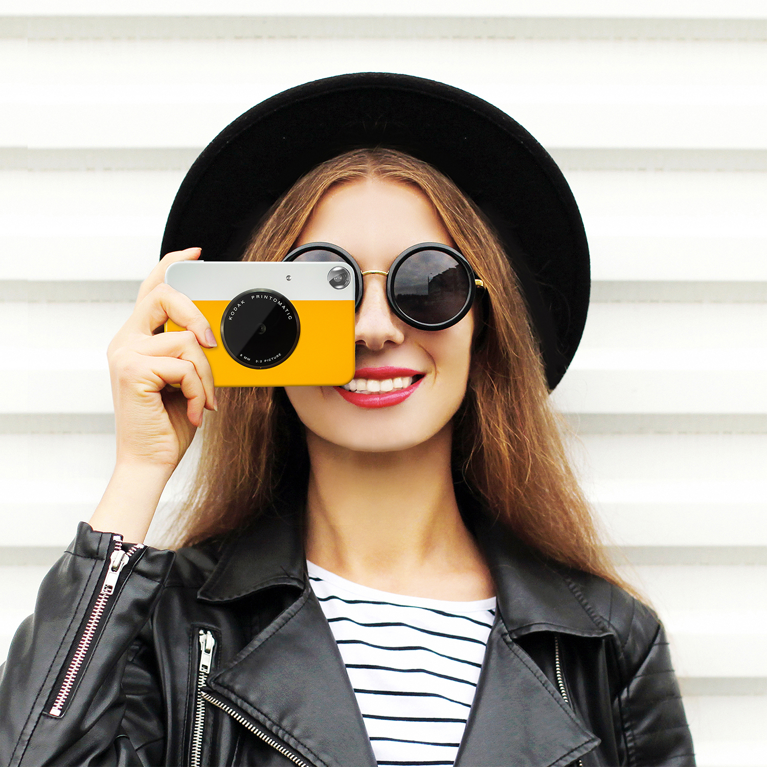 Una donna con una macchina fotografica all'occhio, indossa cappello e occhiali da sole, sorridente.