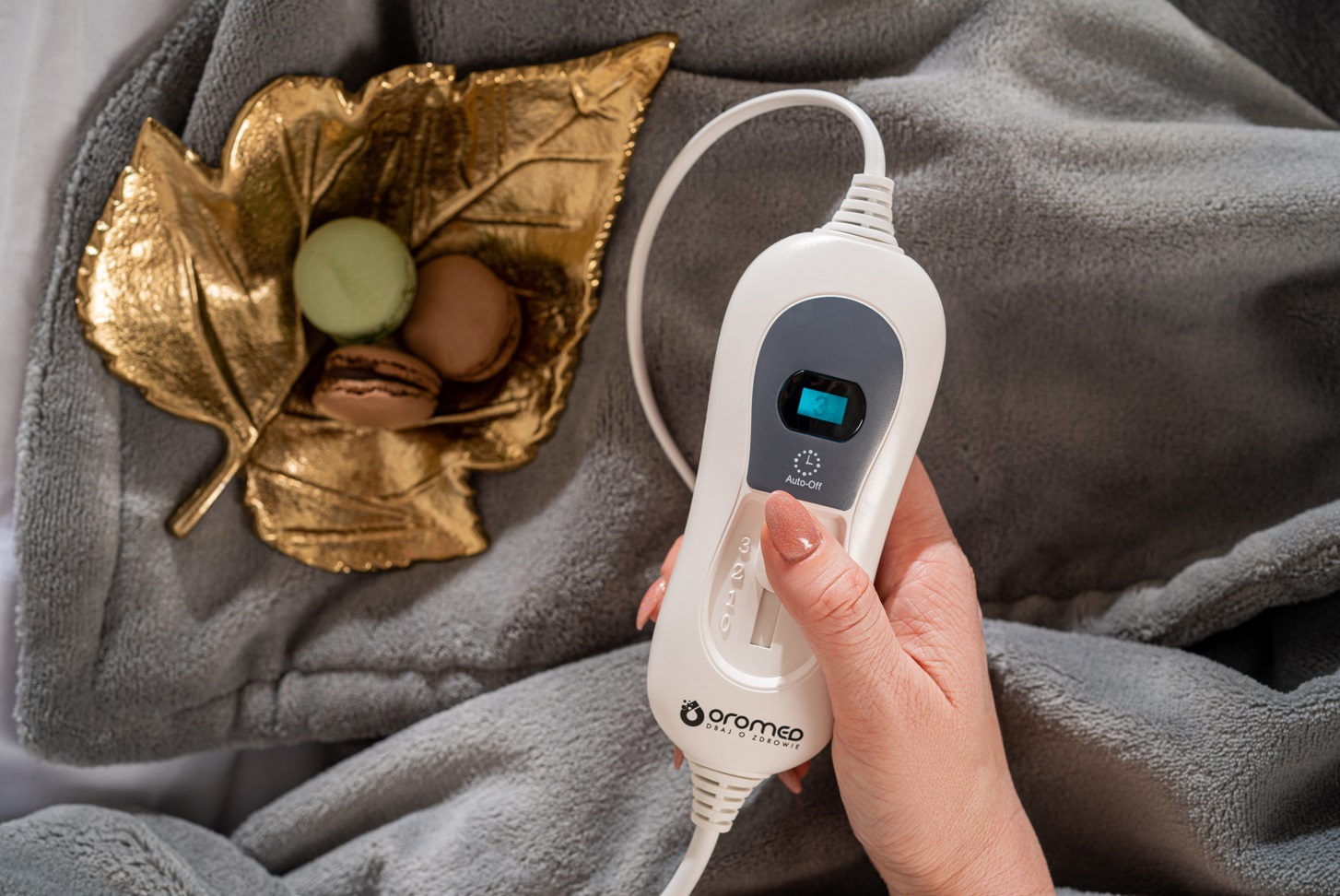 Ręka trzyma panel sterowania do koca grzewczego. W pobliżu złote liście i makaroniki.