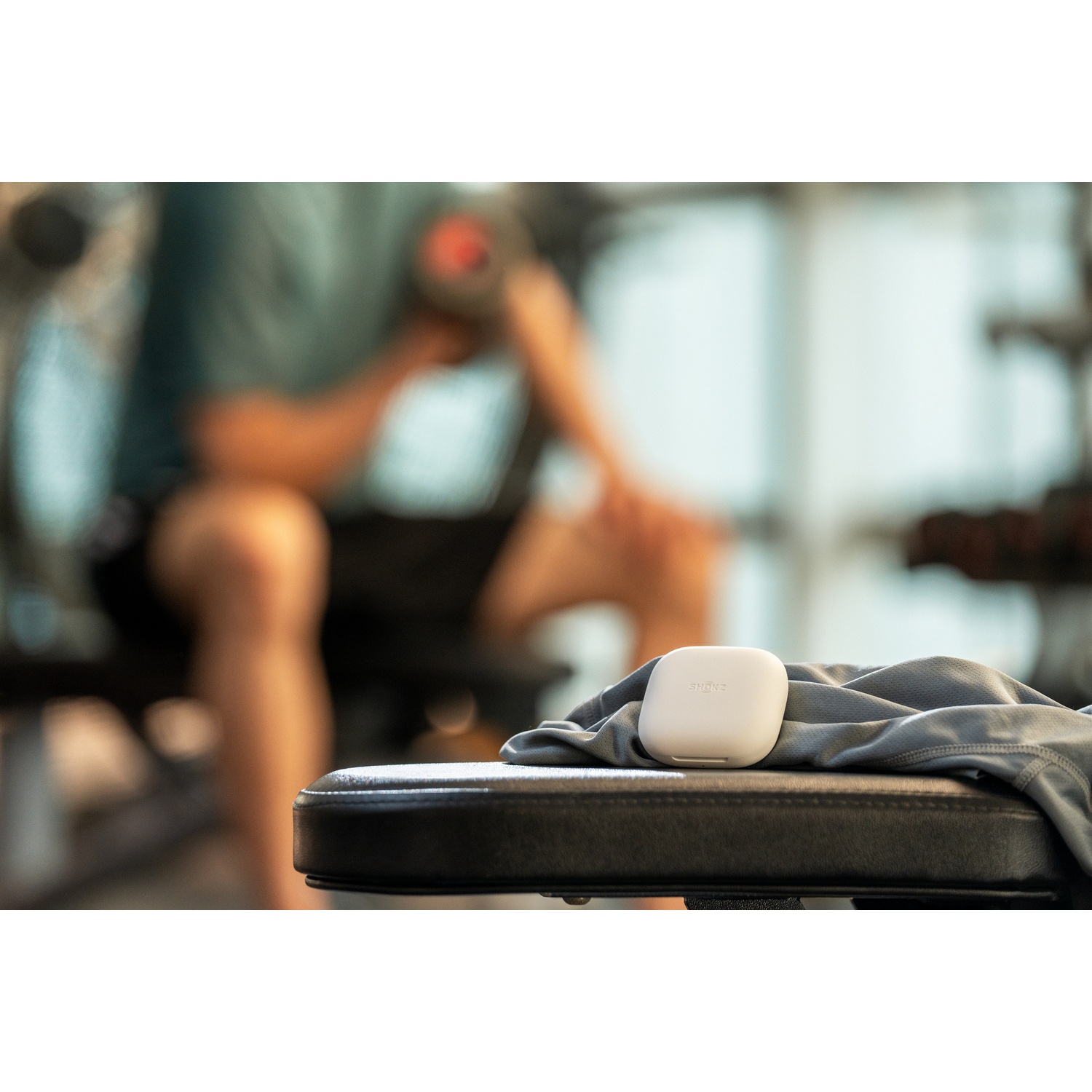 Étui d'écouteurs blanc sur un banc. Un homme flou en arrière-plan, s'exerçant dans une salle de sport.