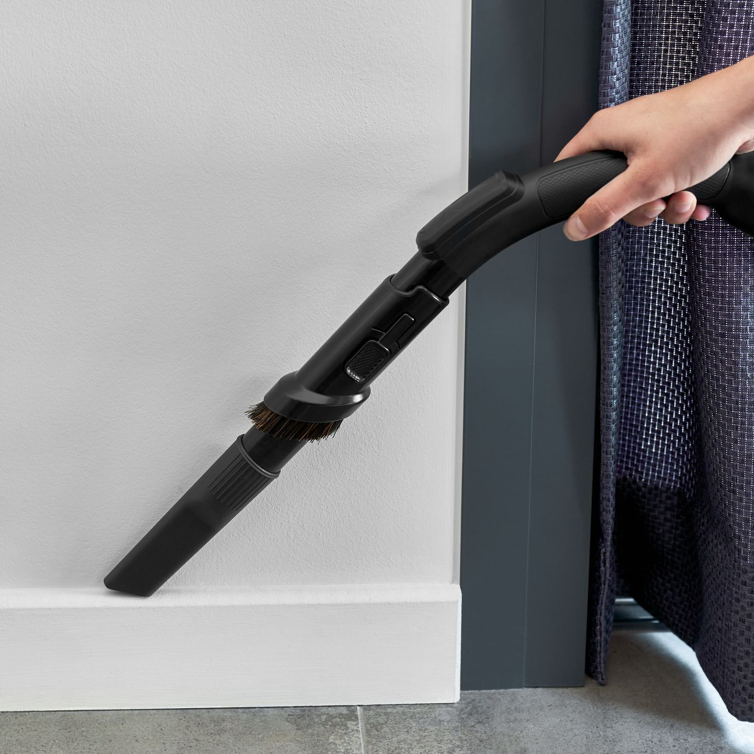 Een hand houdt een zwart stofzuigeropzetstuk vast dat een witte muur schoonmaakt. Het opzetstuk heeft borstels en bevindt zich bij een donkere deur en gordijn.