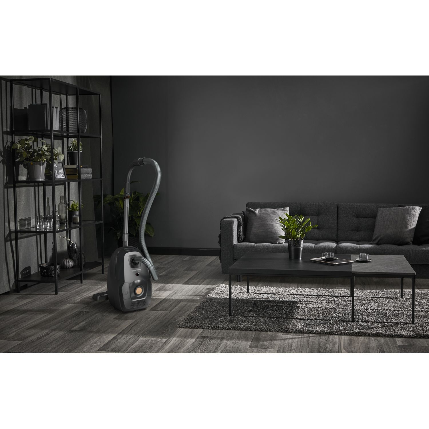 Een moderne woonkamer met een stofzuiger. Een bank, salontafel en boekenrek zijn ook zichtbaar, allemaal in grijstinten.