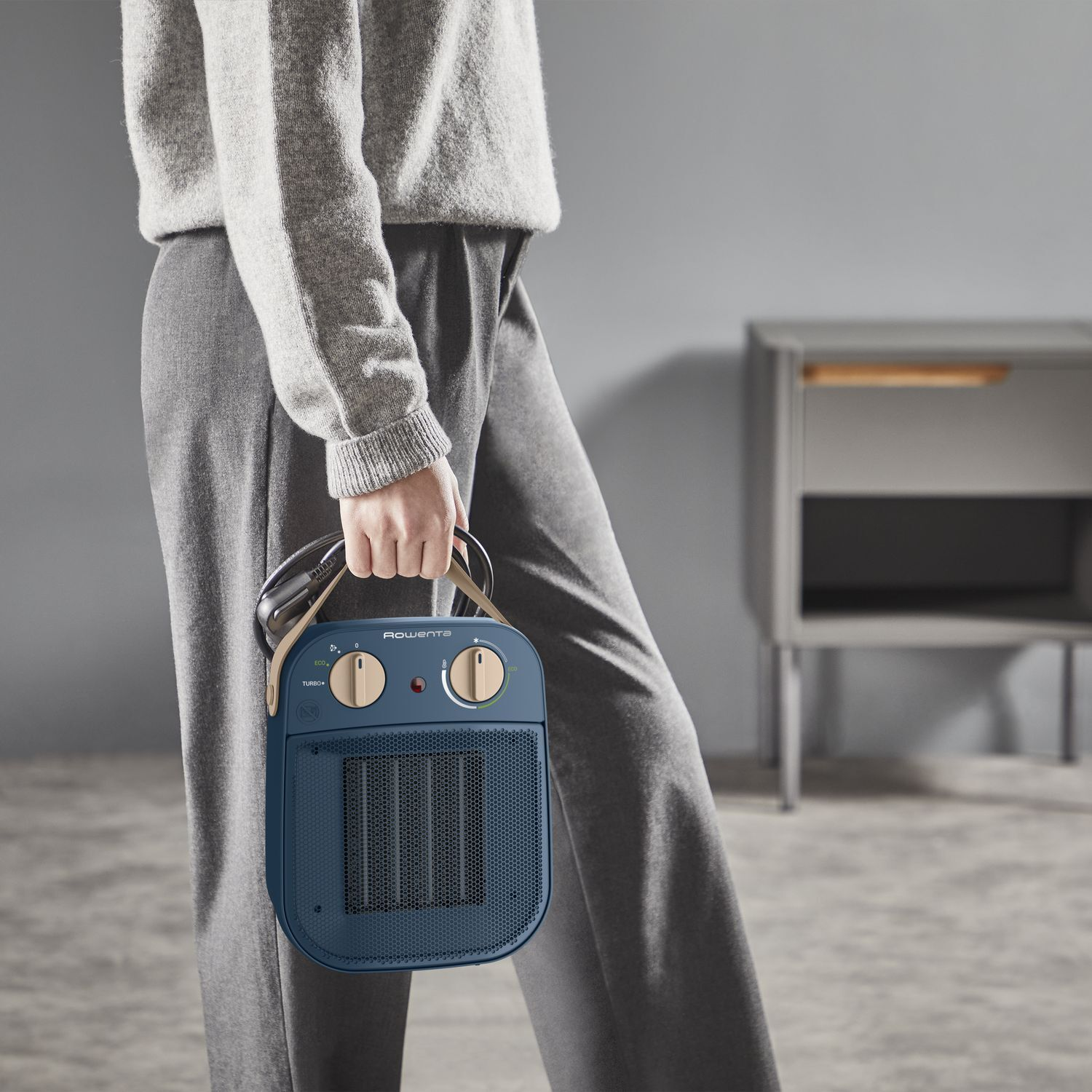 Une personne en vêtements gris tient un radiateur Rowenta bleu et beige. Un meuble gris est à l'arrière-plan.