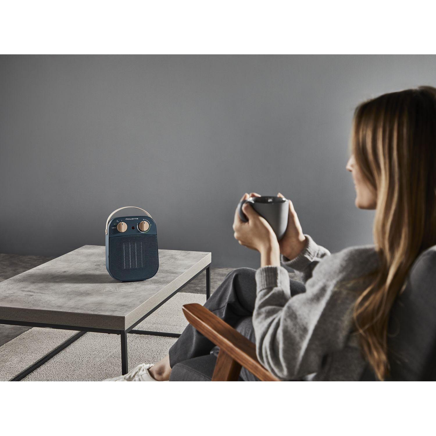 Une femme en pull gris est assise, tenant une tasse. Un radiateur bleu est sur la table. Le fond est un mur gris.