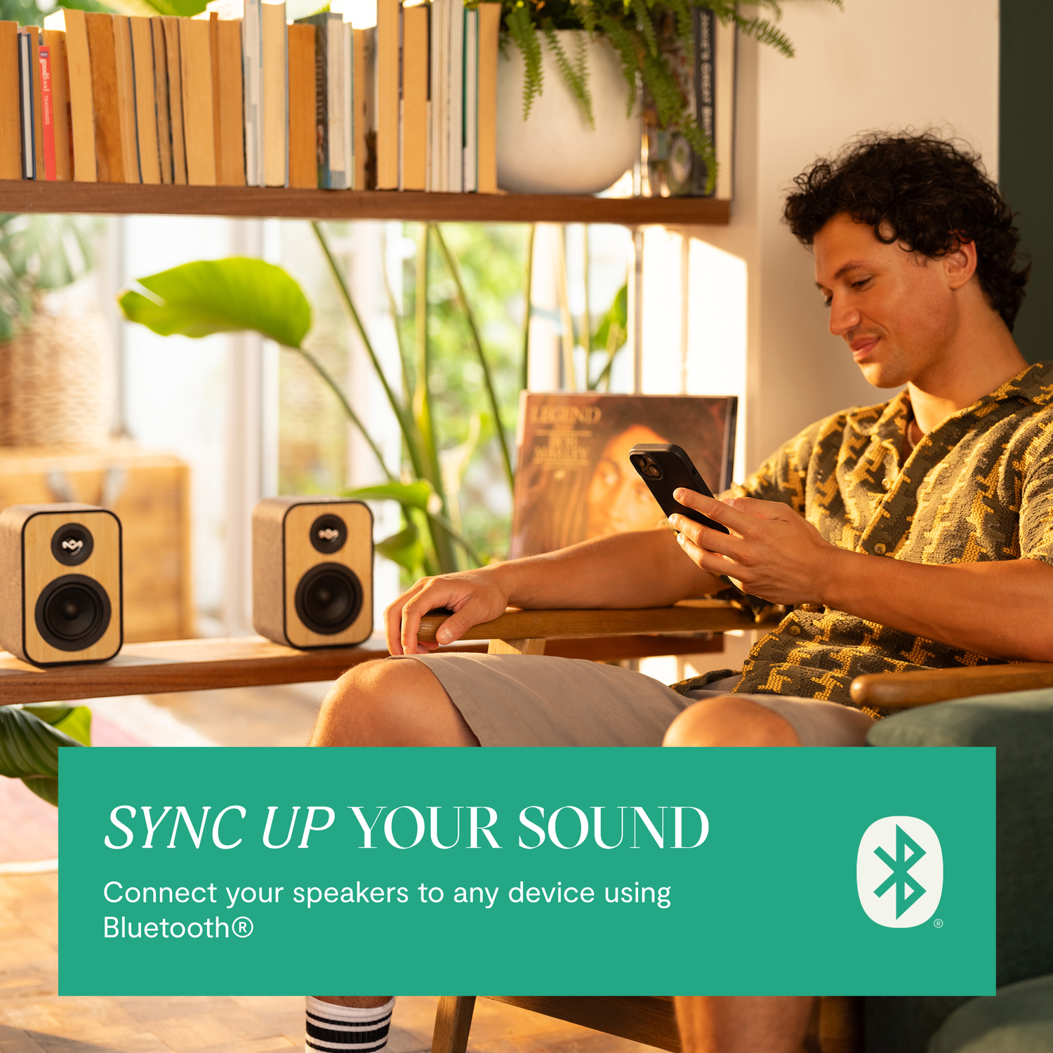 Een man die een telefoon gebruikt terwijl hij in de buurt van twee luidsprekers zit. Tekst zegt 'Sync Up Your Sound'.