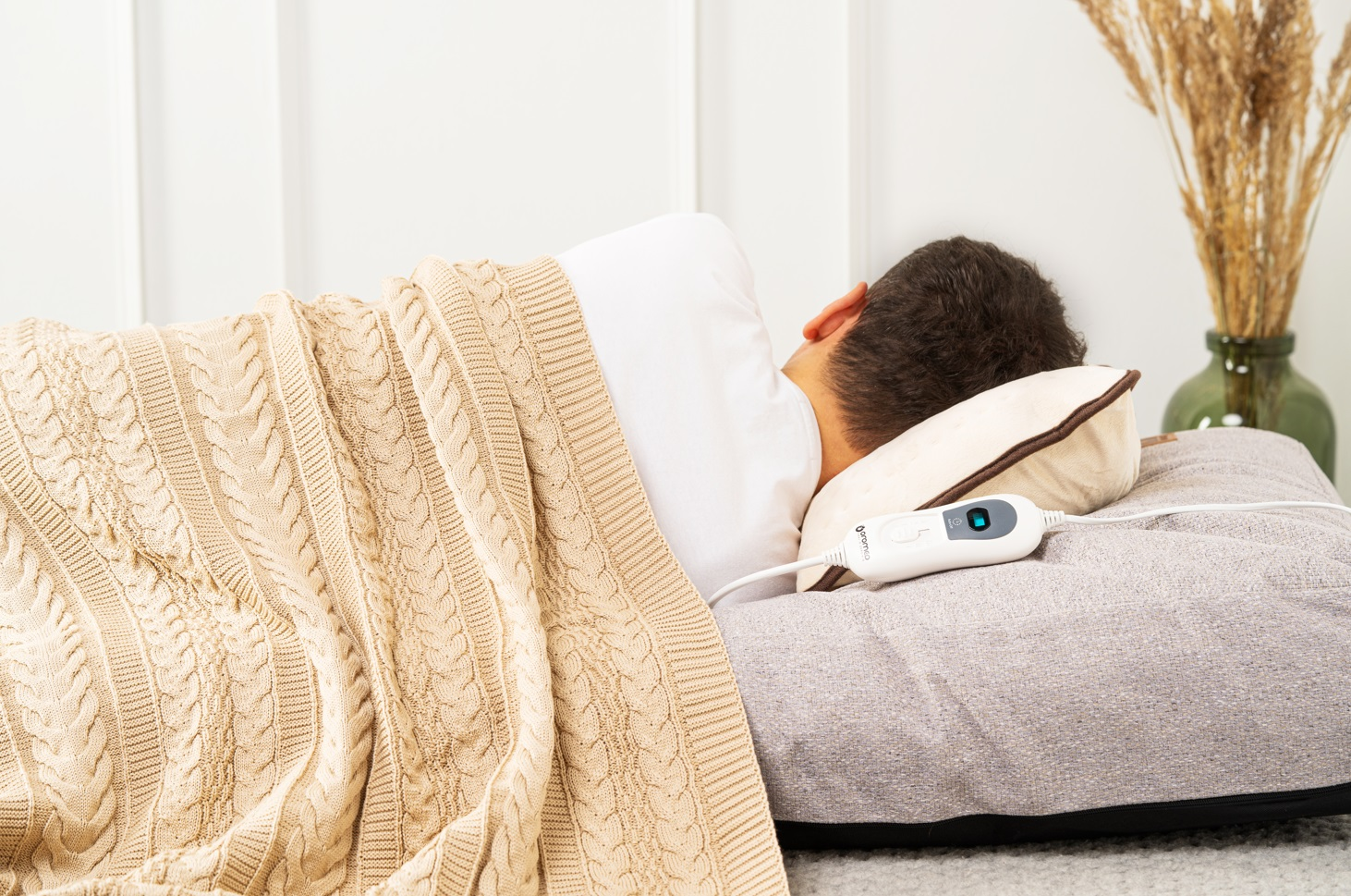 Mężczyzna leżący na łóżku z poduszką rozgrzewającą. Beżowy koc, biała ściana.