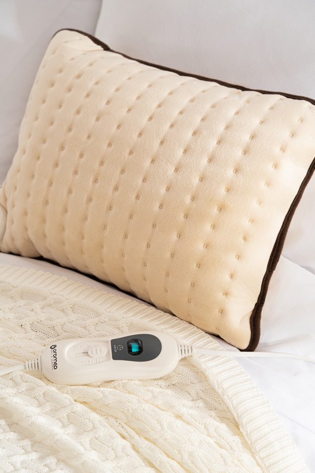 Beżowa poduszka grzewcza z brązową obwódką, na białym łóżku.