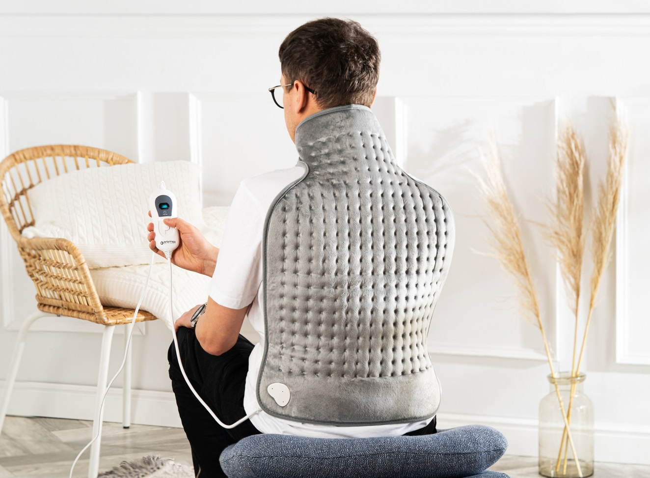 Mężczyzna nakłada poduszkę grzewczą na plecy. Jest w białym pokoju. Widoczna jest drabina i roślina.