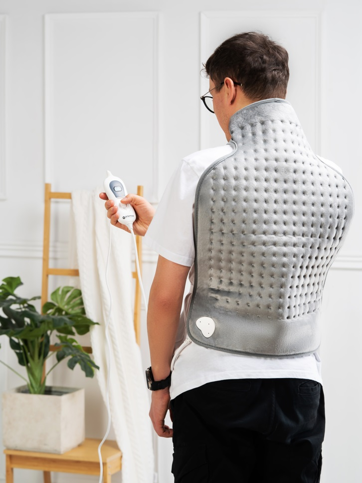 Mężczyzna nakłada poduszkę grzewczą na plecy. Jest w białym pokoju. Widoczna jest drabina i roślina.