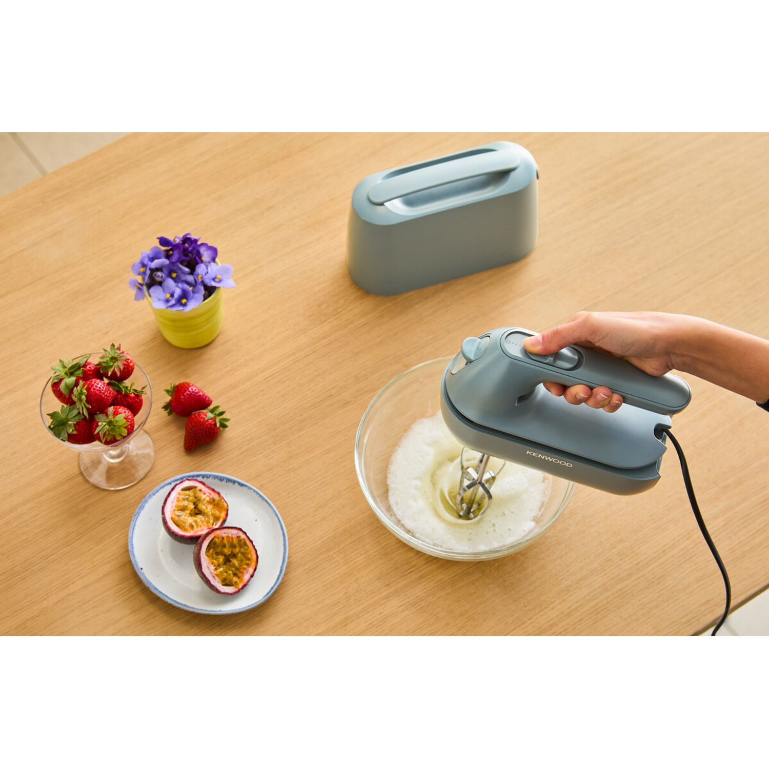 A person is using a gray hand mixer. There are strawberries, flowers, and passion fruit.