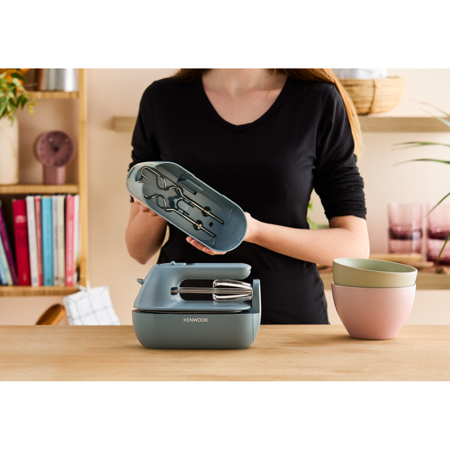 A woman holding a mixer with beaters. Two bowls sit on the wooden table.
