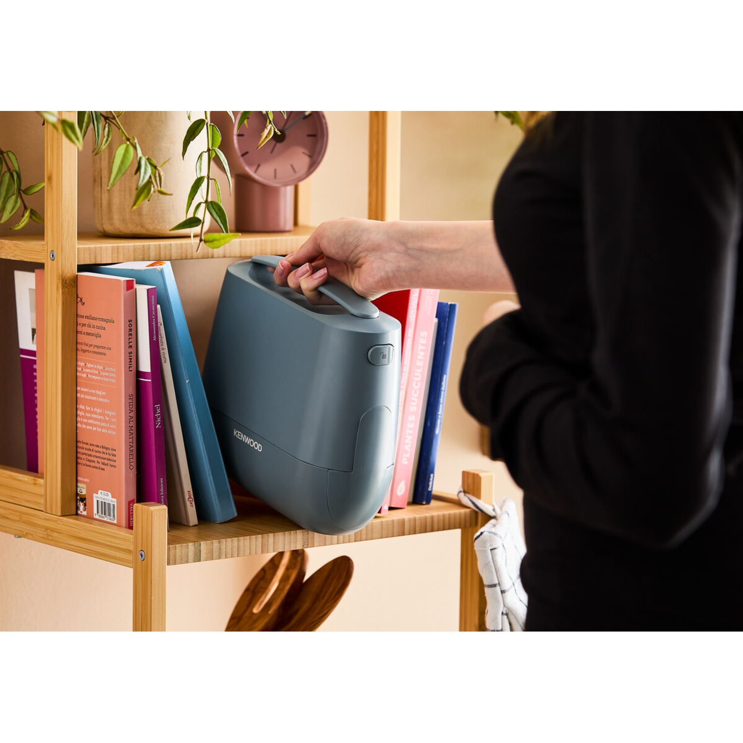 Woman holding a grey Kenwood device near books on wooden shelves.