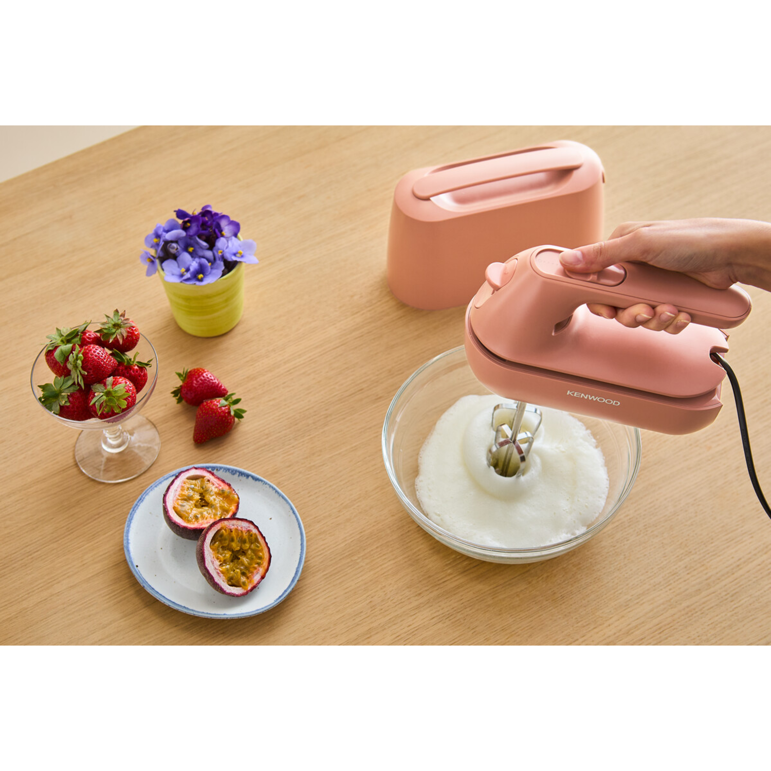 Person using a pink hand mixer in a bowl with whipped cream, and fruits.