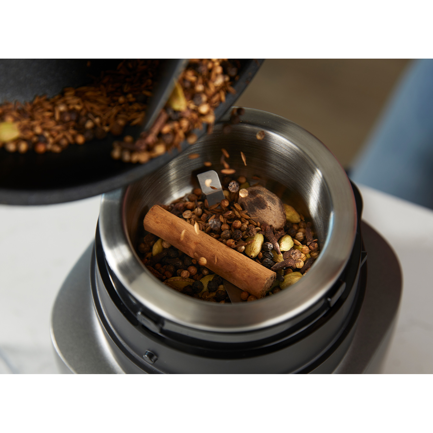Spice grinder with spices being poured in. Cinnamon stick and other spices are visible.
