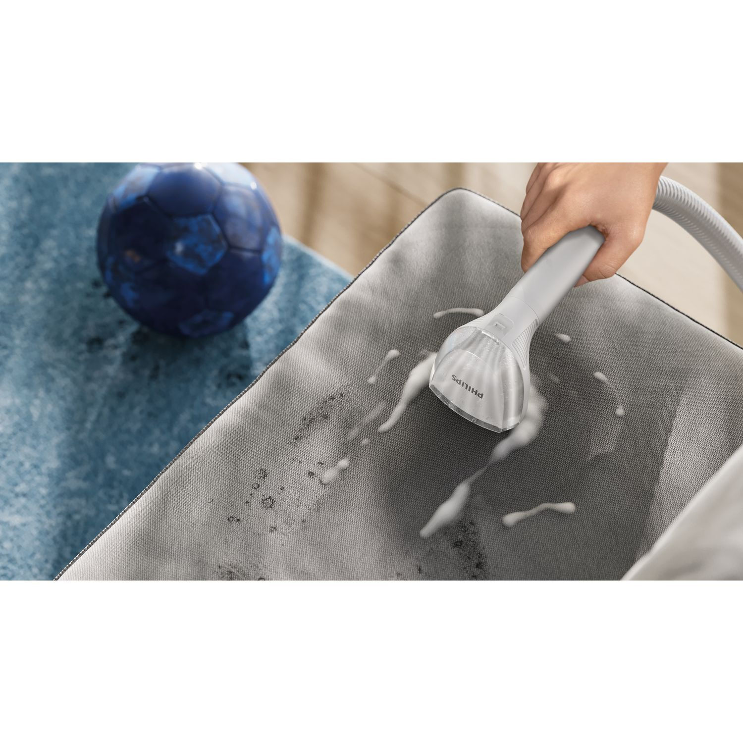 Une main vaporise un fauteuil en tissu gris avec de la mousse blanche. Une balle bleue est à gauche.