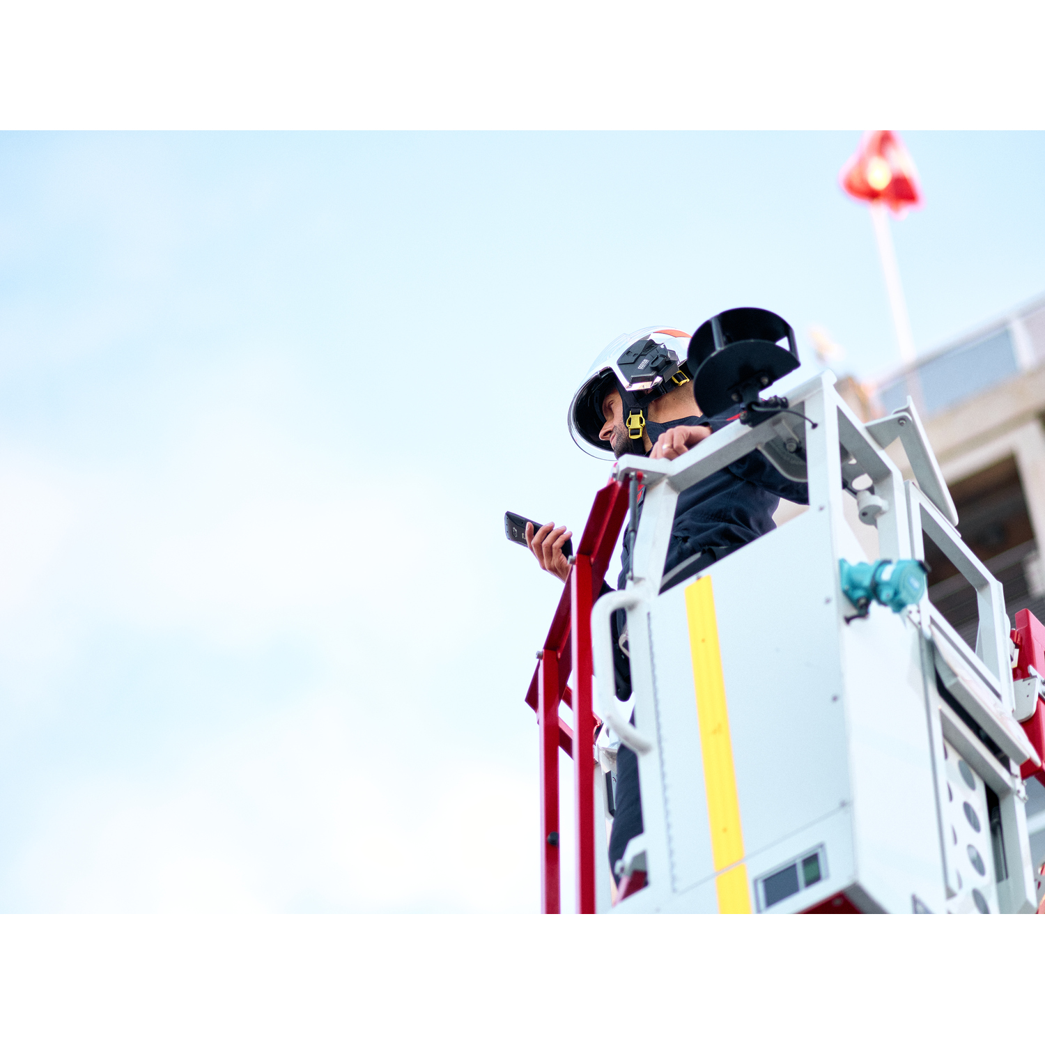 Brandweerman in een lift, opzij kijkend, een telefoon vasthoudend, blauwe lucht op de achtergrond.
