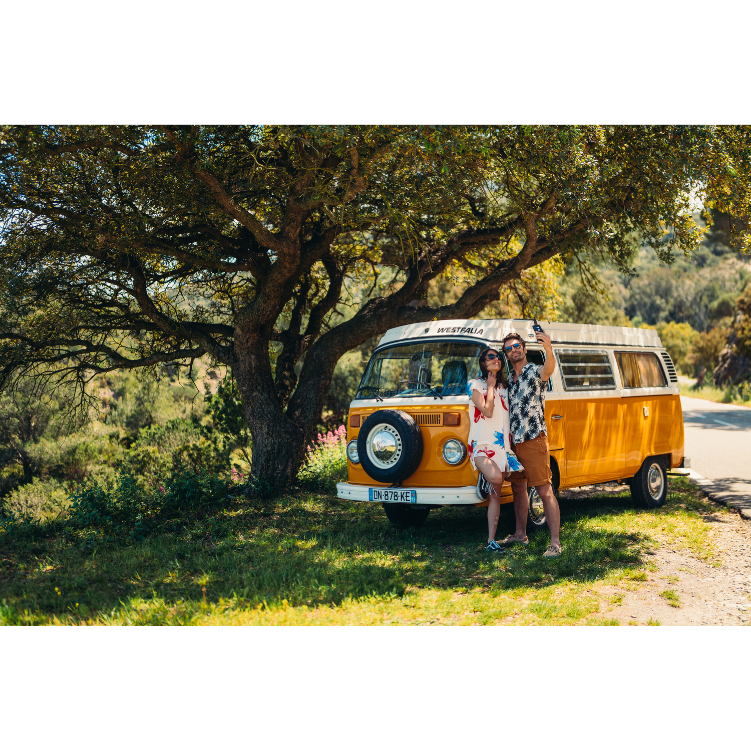 Een stel maakt een selfie naast een gele VW-camper onder een boom.