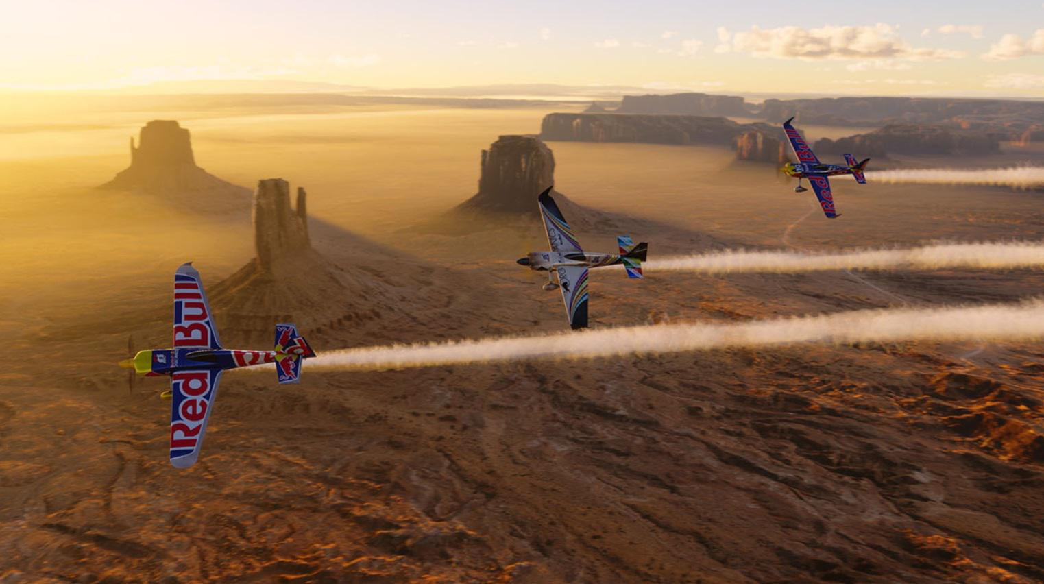 Drei Red Bull-Flugzeuge fliegen in einer Wüstenlandschaft. Flugzeuge hinterlassen weiße Kondensstreifen.