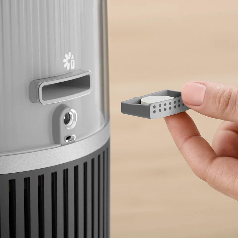 Ręka trzyma małe, szare urządzenie z małą białą pigułką. Urządzenie ma otwory.