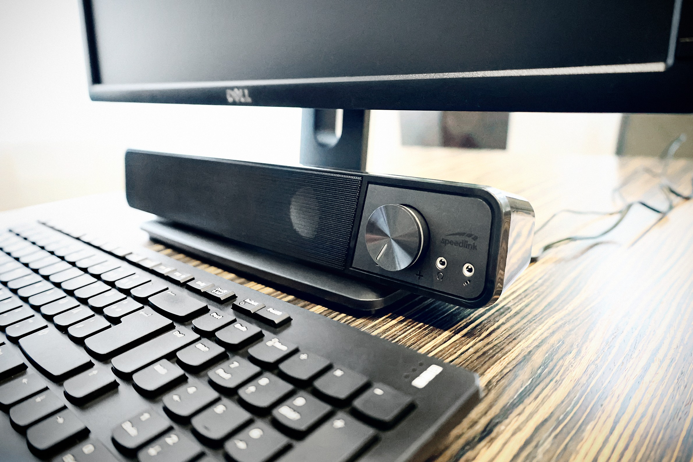 Eine schwarze Soundbar vor einem Computermonitor mit Tastatur auf einem Holzschreibtisch.