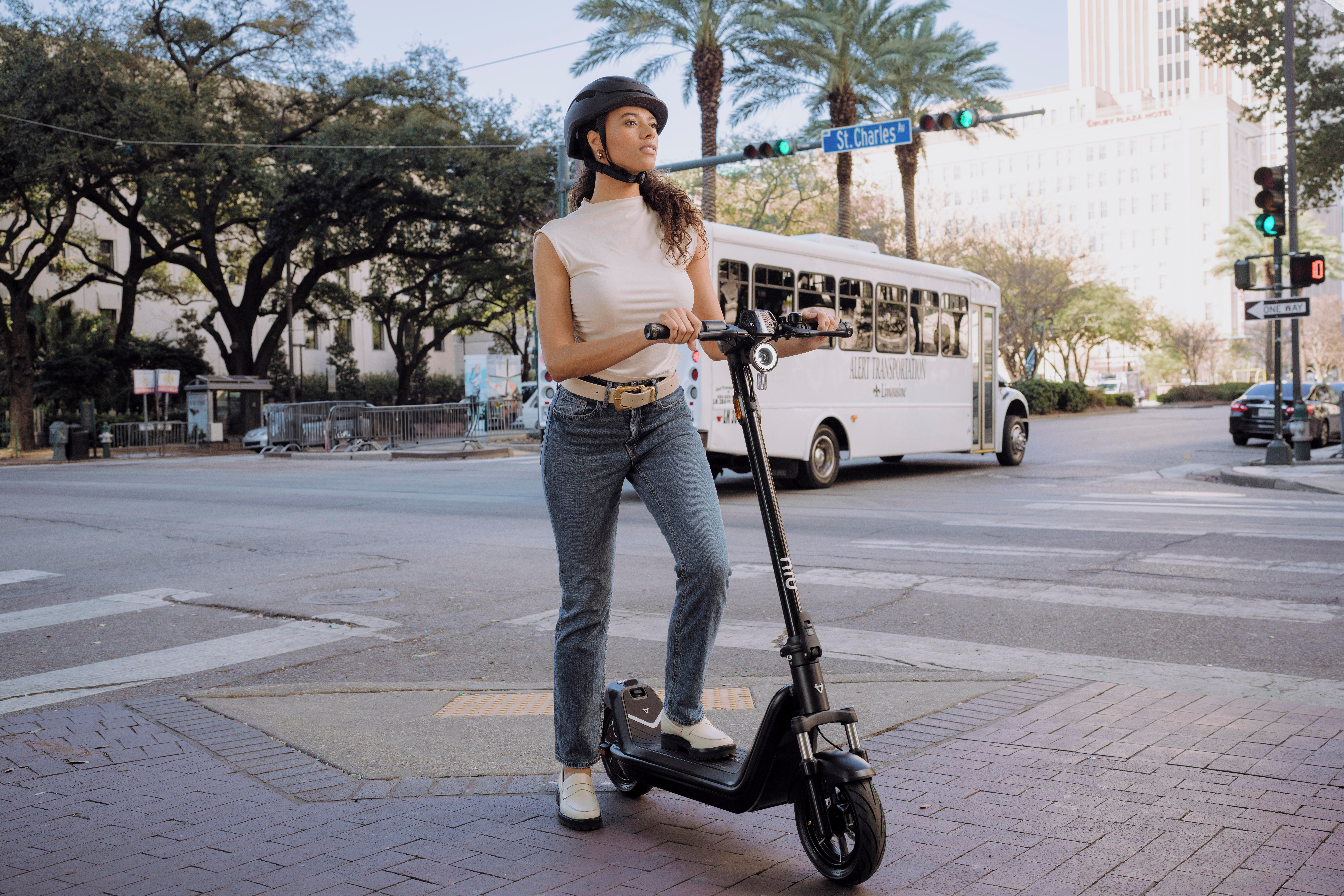 Frau auf Roller auf der Straße, Helm tragend. Gebäude und Bäume im Hintergrund.